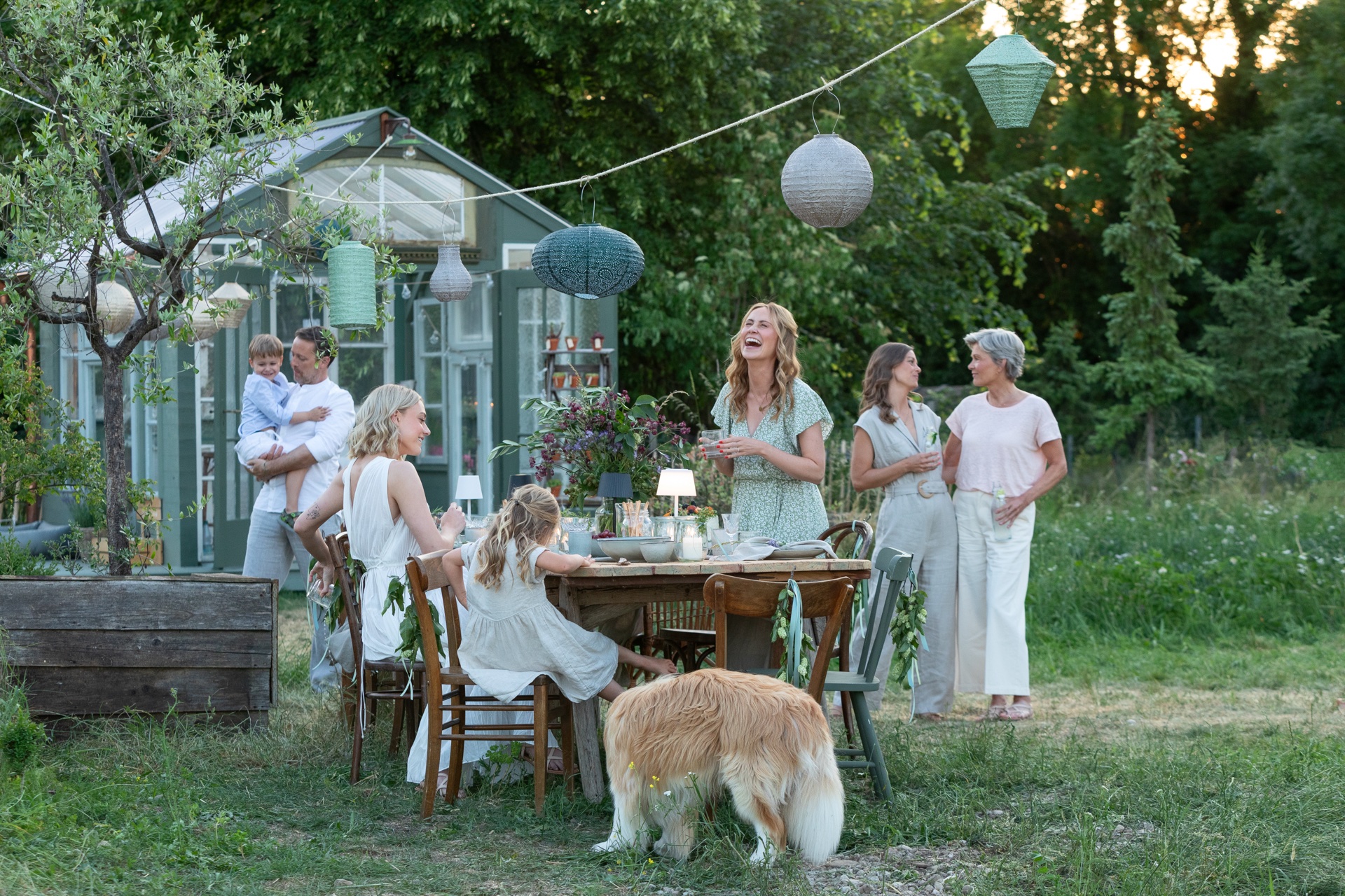 Familie am gedeckten Tisch unter Lampions vor dem mintgrünen Gewächshaus am Abend