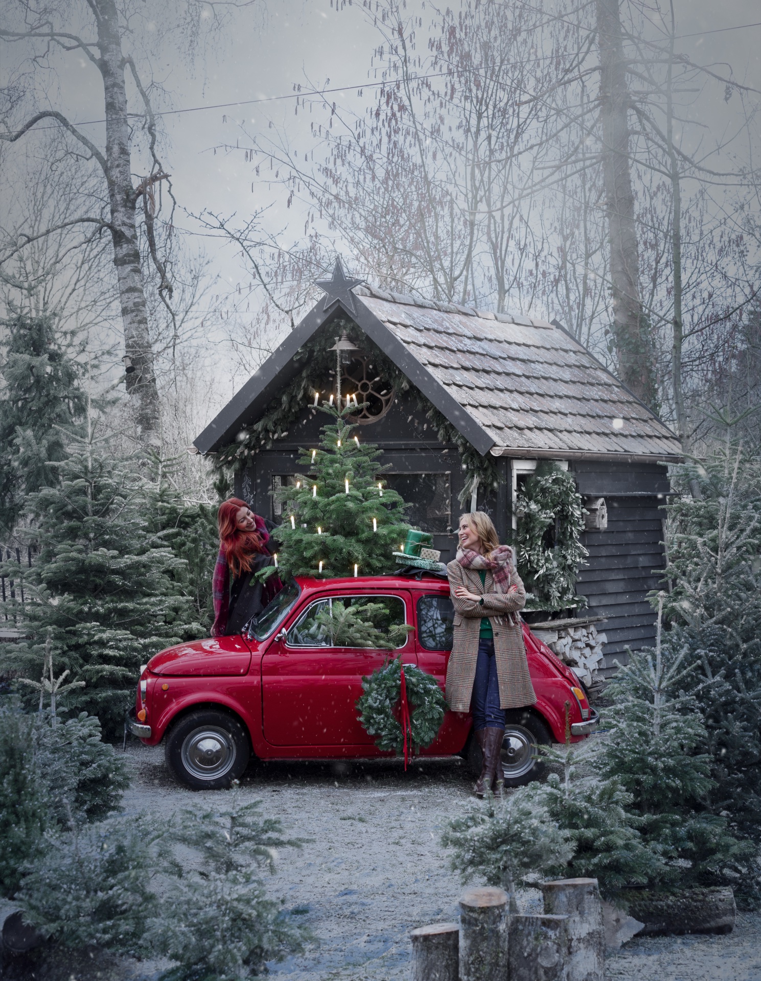 Roter Fiat 500 mit Weihnachtskranz und kleinem Tannenbaum auf dem Dach vor schwarzer Hütte im verschneiten Wald